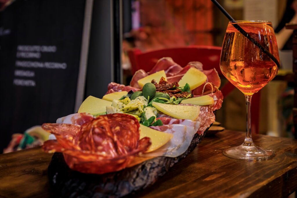 Aperol Spritz cocktail with ice and a straw next to a rustic Italian charcuterie board featuring cured meats, cheeses, olives, and artichokes on a wooden table.